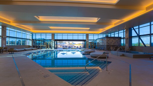 Piscina interior moderna con paredes de vidrio transparente, terraza con mobiliario y vistas al exterior.