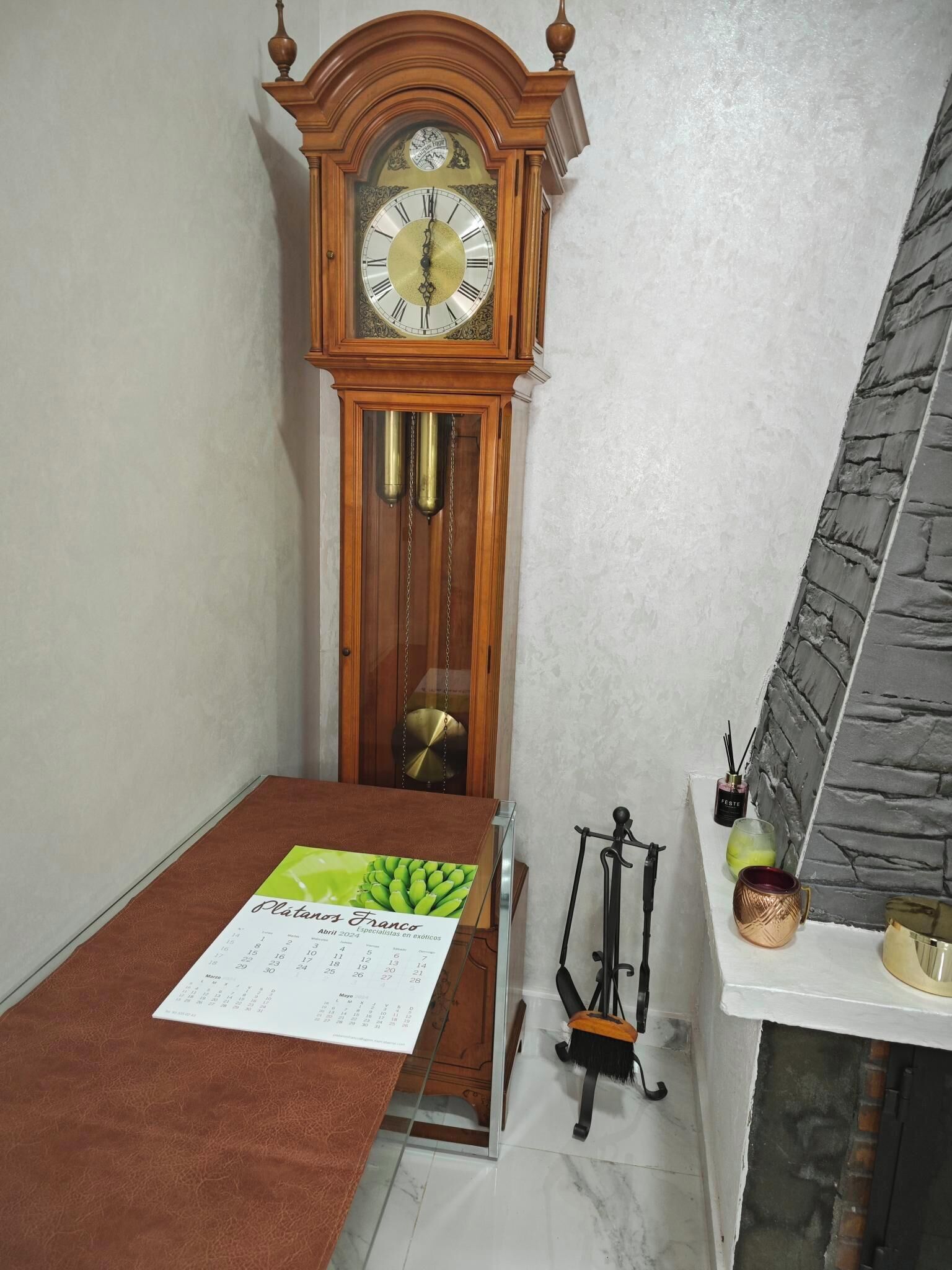 A room with a wooden grandfather clock, a calendar on a table, a small shelf with decorative items, and a stone-textured wall.