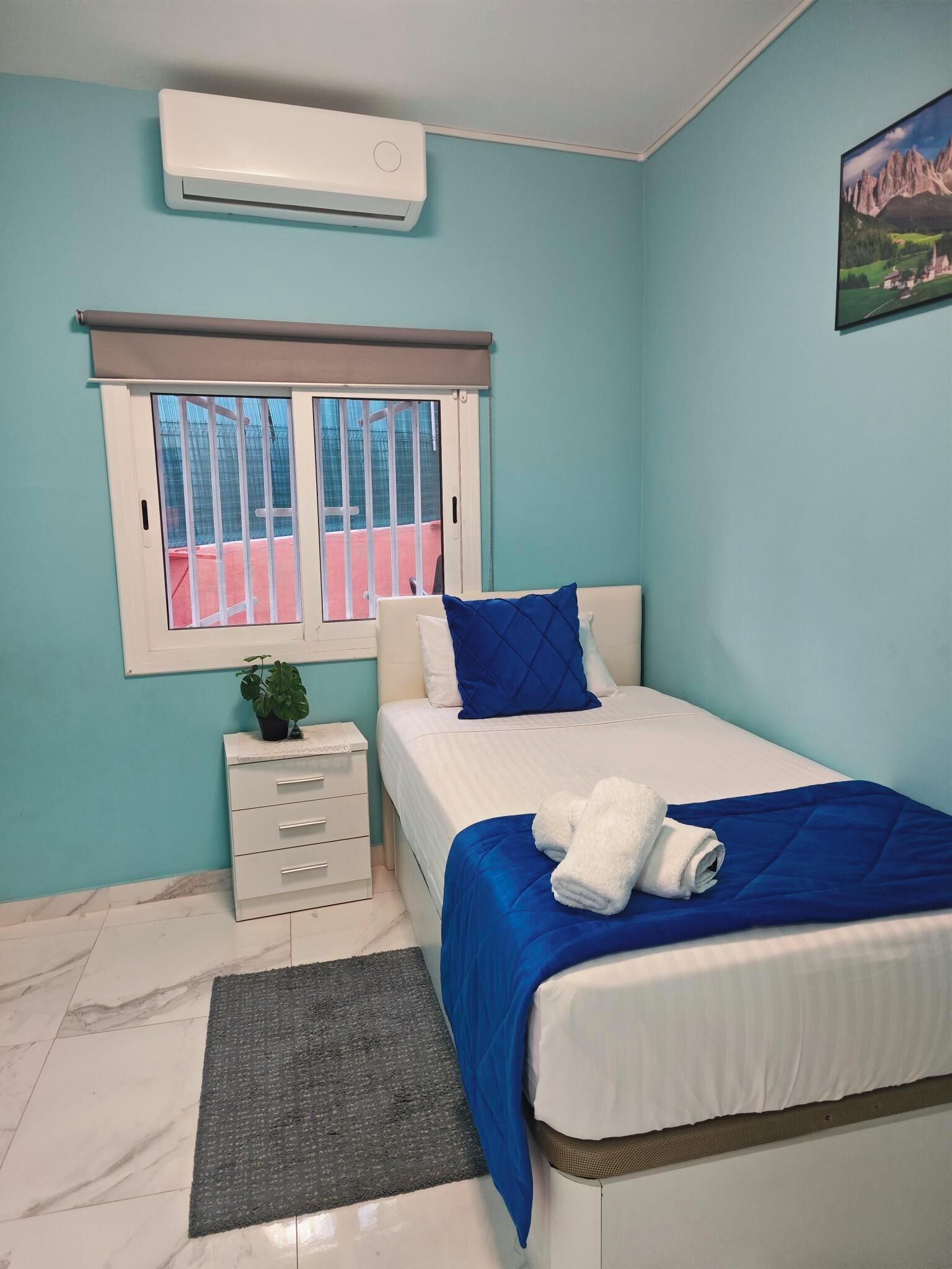 Un dormitorio con cama, ventana con vistas, una mesita de noche pequeña y aire acondicionado instalado en la pared.
