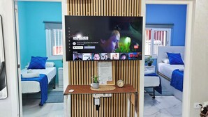 A hotel room with a large flat-screen TV mounted on a wooden panel wall, a bed with blue bedding, a small bedside table with a plant, and a white air conditioning unit above the TV. - RELAX HOUSE BARCELONA (Sant Esteve Sesrovires)