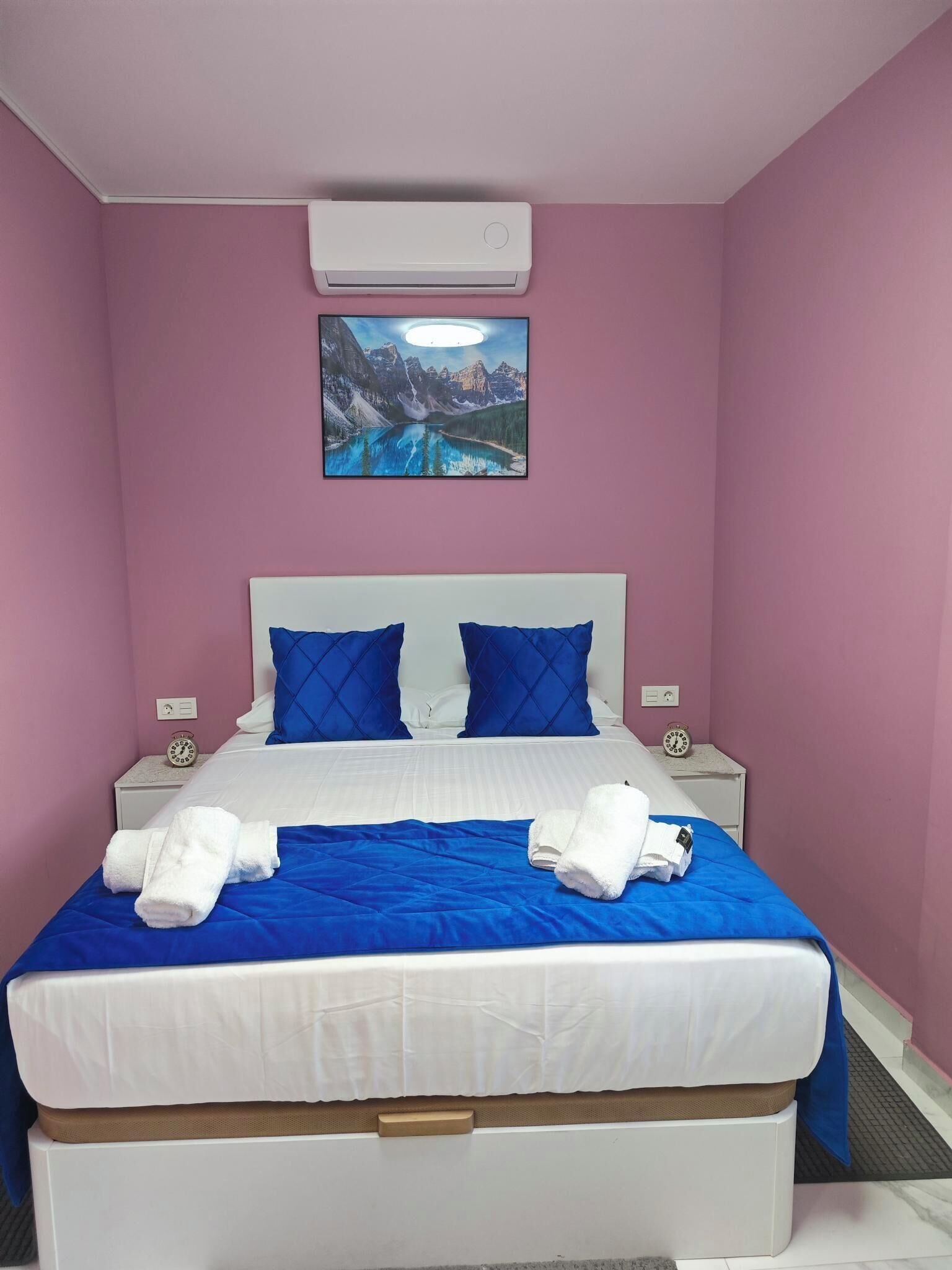 A neatly made bed with blue and white bedding, two pillows, and two white towels on the bed. There is a clock on each bedside table, and a painting of a mountain landscape hangs above the bed.