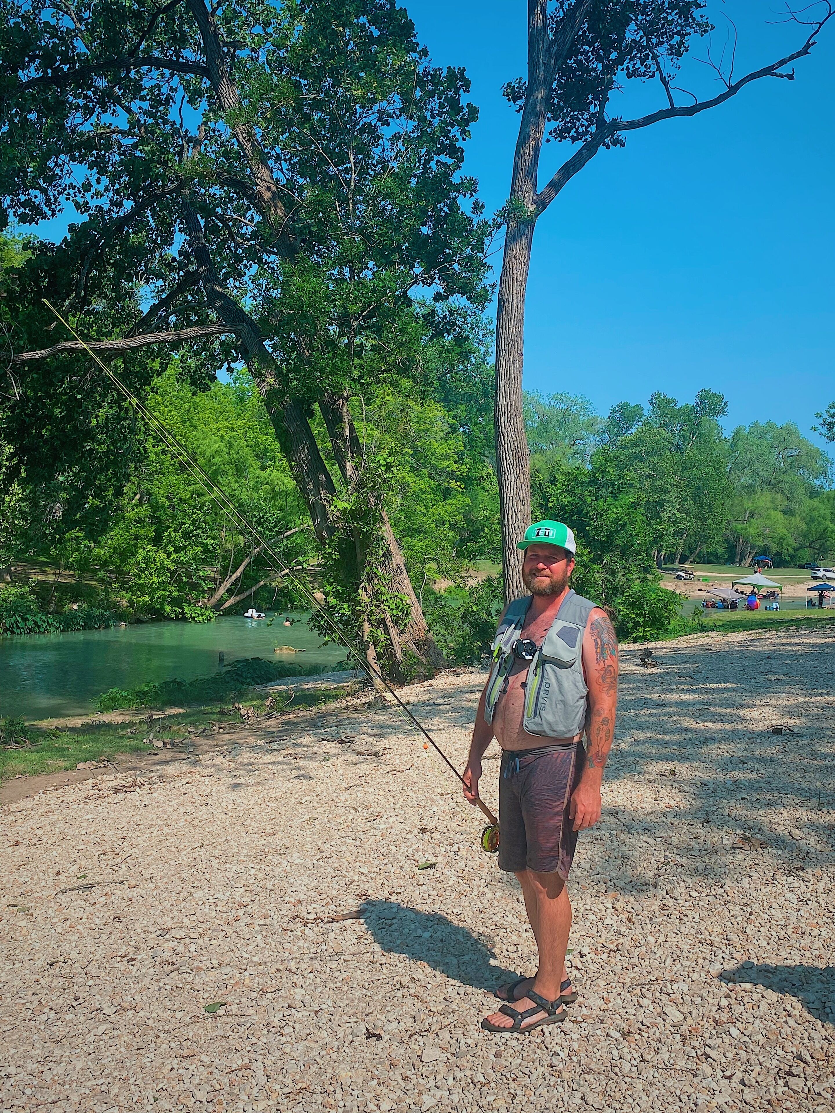 A person standing on a gravel area near a body of water with trees in the background.