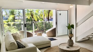 A modern living room with a sofa, wicker chairs, a round coffee table, and a view of a tropical garden through large glass doors.