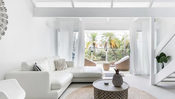 A modern living room with a white sofa, a round coffee table, and a view of a balcony with palm trees.