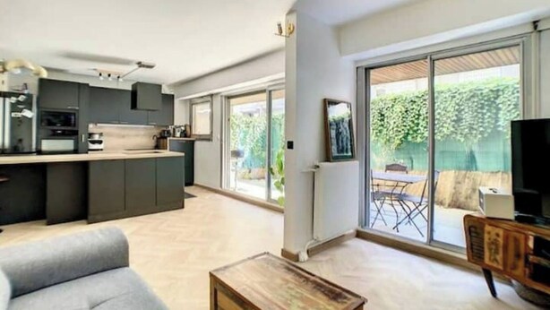 Une cuisine moderne avec des meubles de couleur sombre, un coin repas avec un canapé gris et une grande porte-fenêtre coulissante donnant sur une terrasse.
