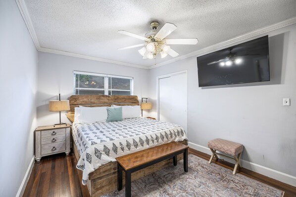 A bedroom with a bed, bedside lamps, a bench, a ceiling fan, a TV, and a window.
