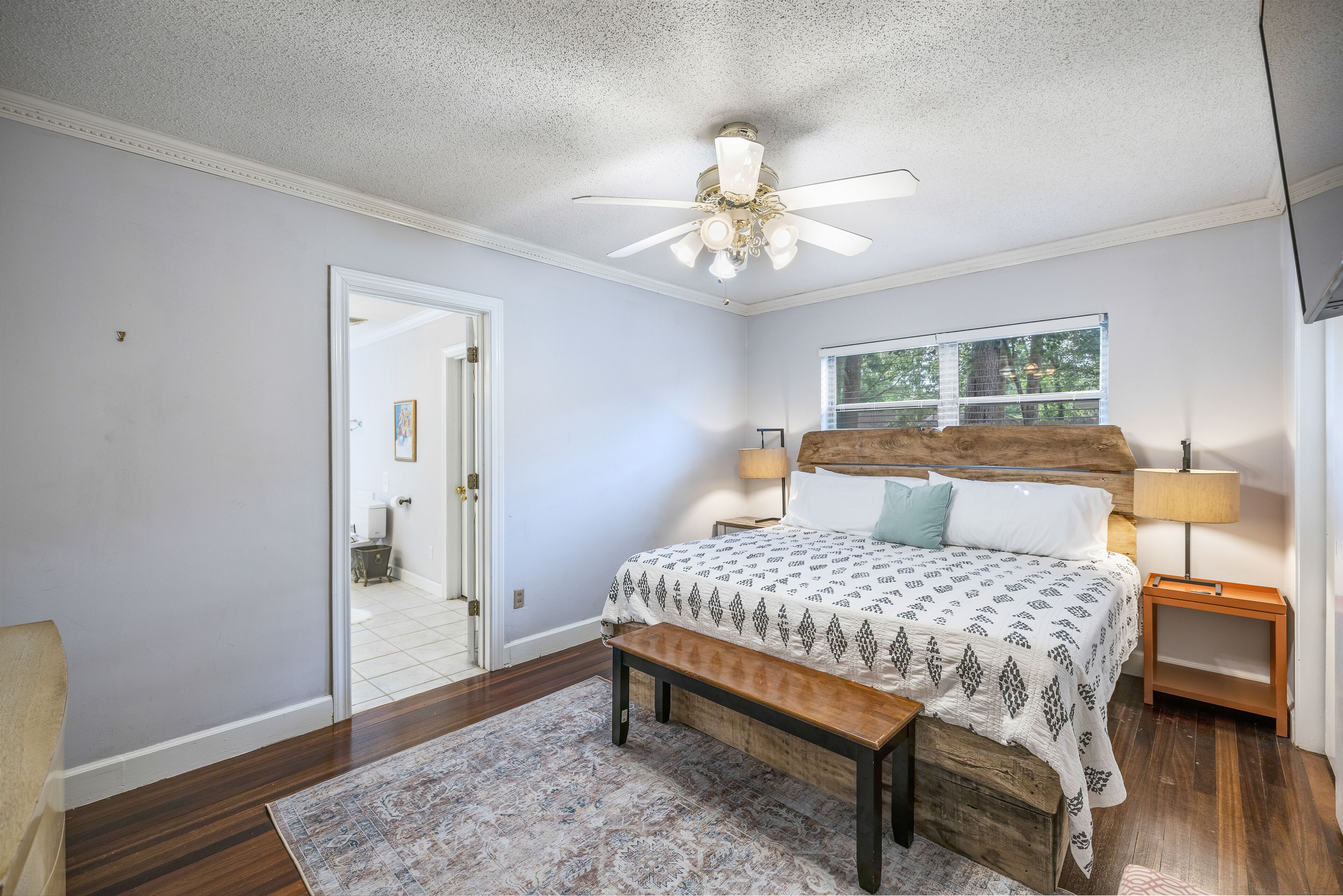 A bedroom with a bed, wooden headboard, two bedside lamps, a ceiling fan, a bench, a rug, and a window with a view of trees.
