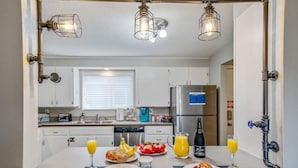 A modern kitchen with white cabinets, a stainless steel refrigerator, and a breakfast setup with bread, fruit, and drinks. - The Industrial - Pet Friendly Home with Garage by Boise Fairgrounds (Boise)