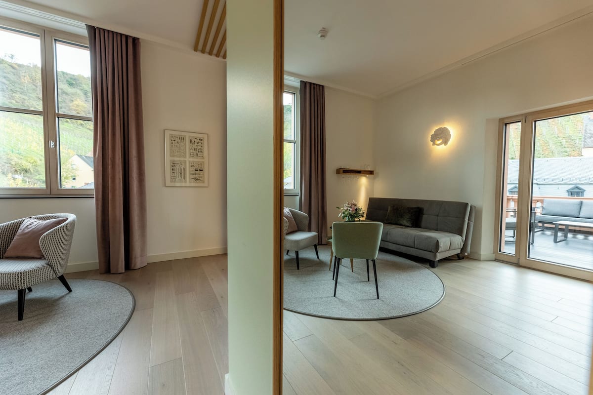 Ein großzügiges Wohnzimmer mit einer Couch, Sesseln, einem Couchtisch und einem großen Fenster mit Vorhängen.