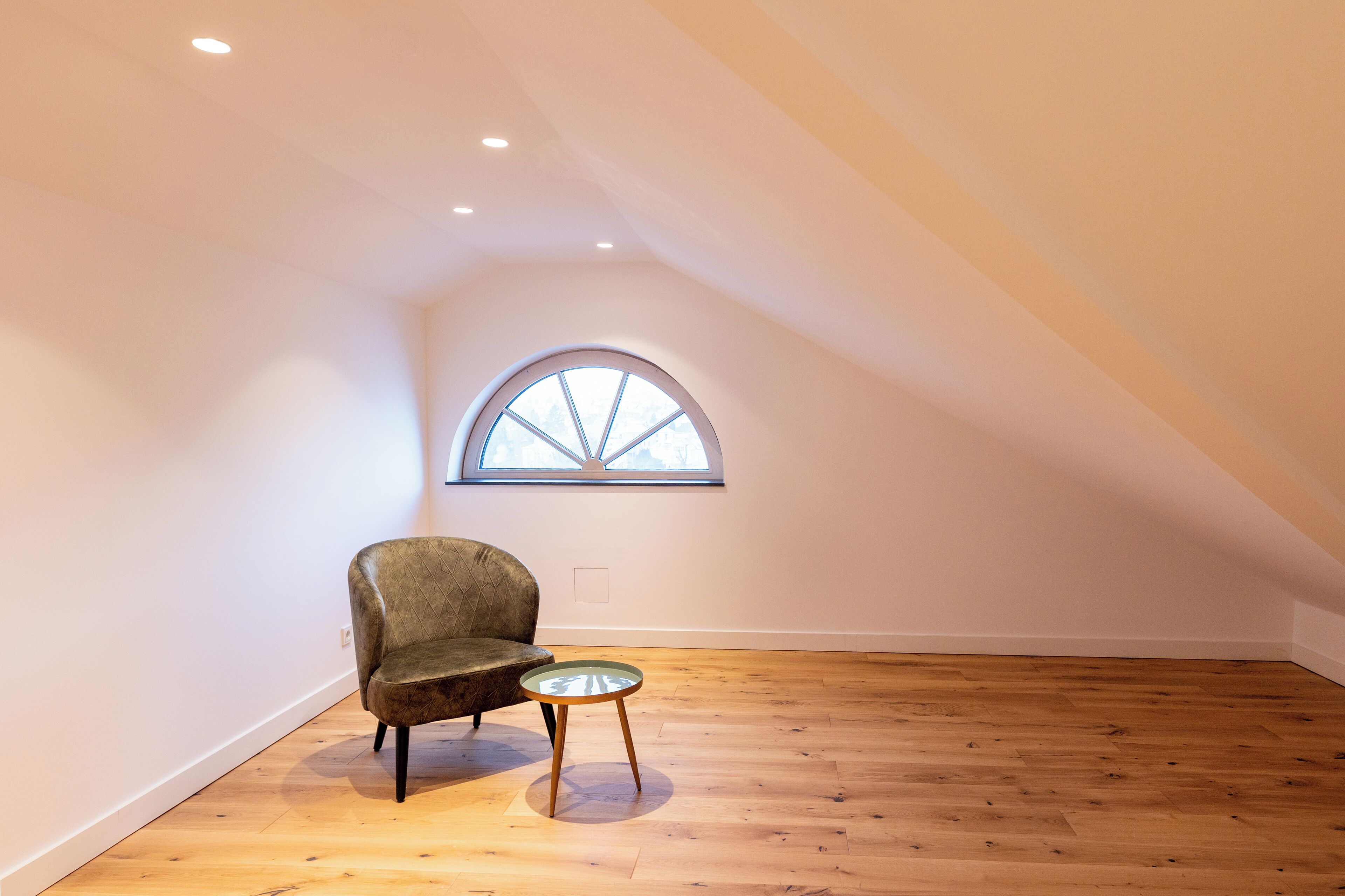 Une pièce mansardée avec un plancher en bois, une fenêtre cintrée, une chaise et une petite table.