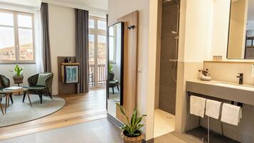 A modern bathroom with a shower, a sink, and a mirror. There is a plant on the floor and a chair in the adjacent living area.