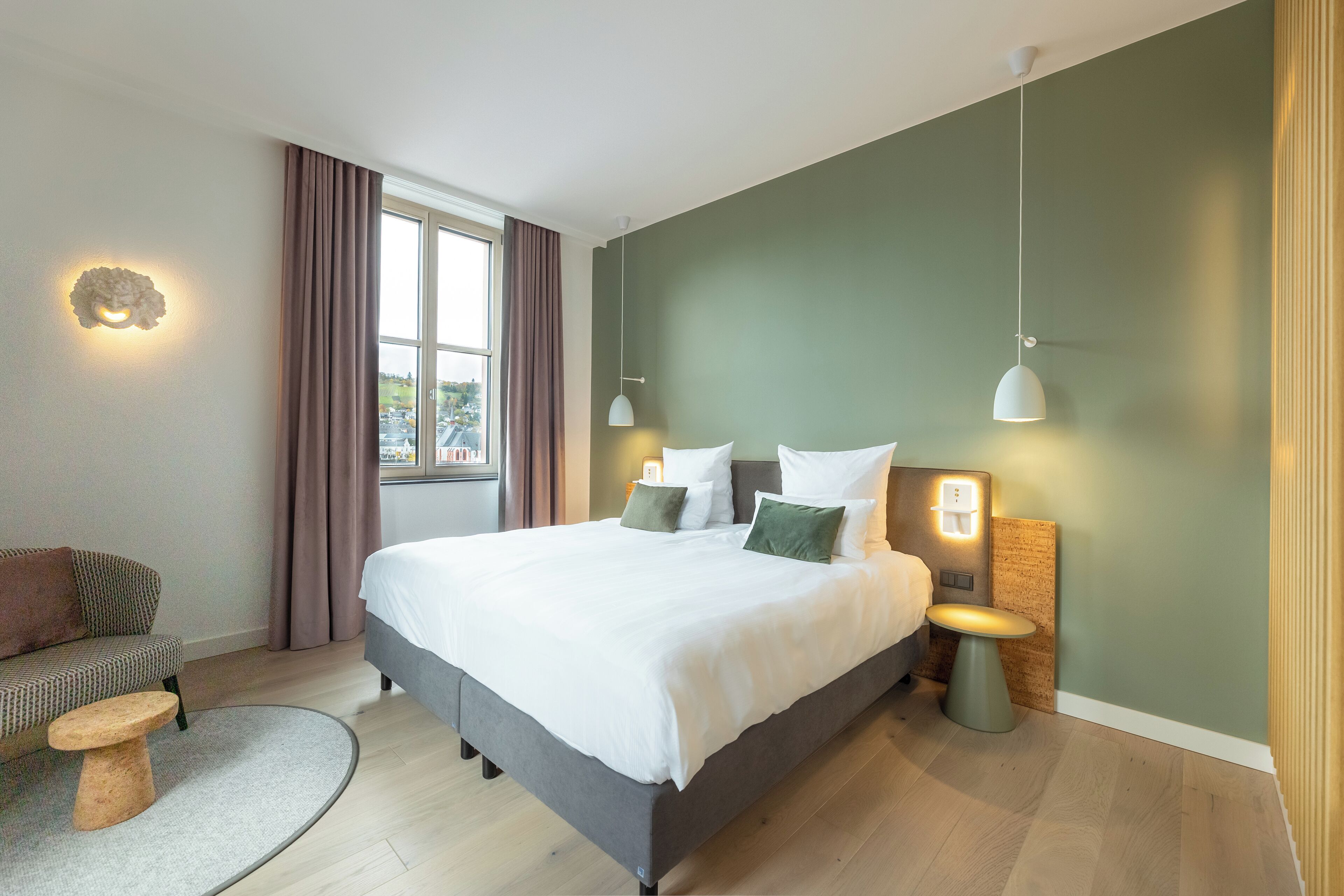 Una habitación de hotel moderna con una cama grande, un sofá, una mesita de centro redonda y vistas a la ciudad desde la ventana.