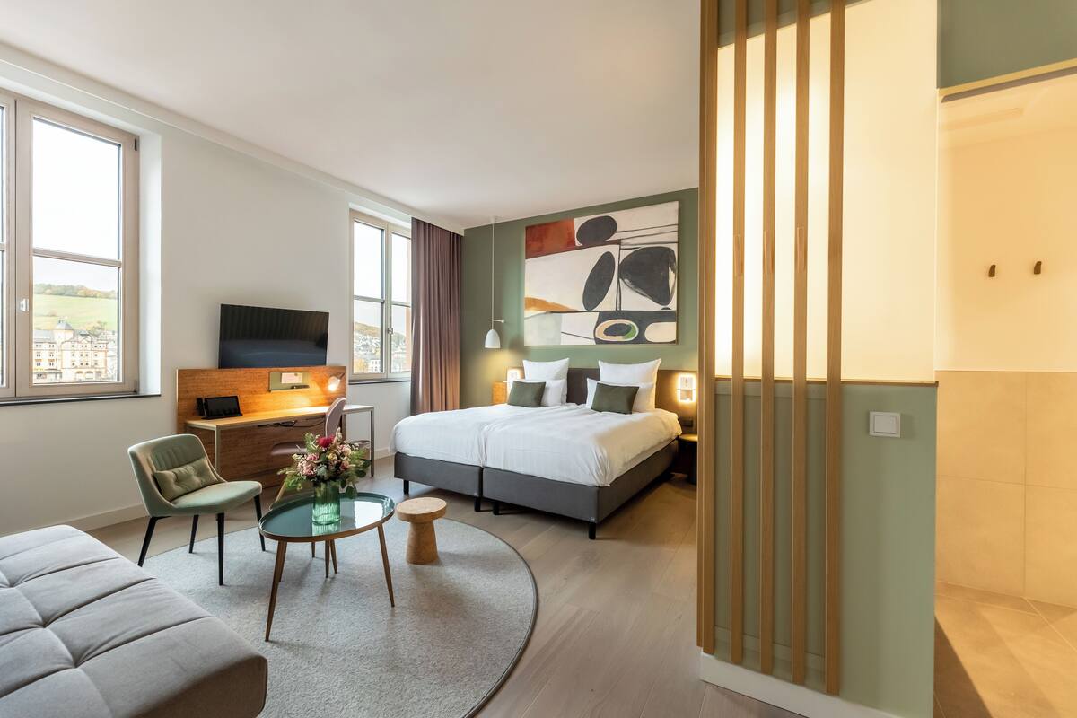 Ein modernes Hotelzimmer mit Bett, Schreibtisch, Fernseher, Stuhl und Blick ins Grüne.