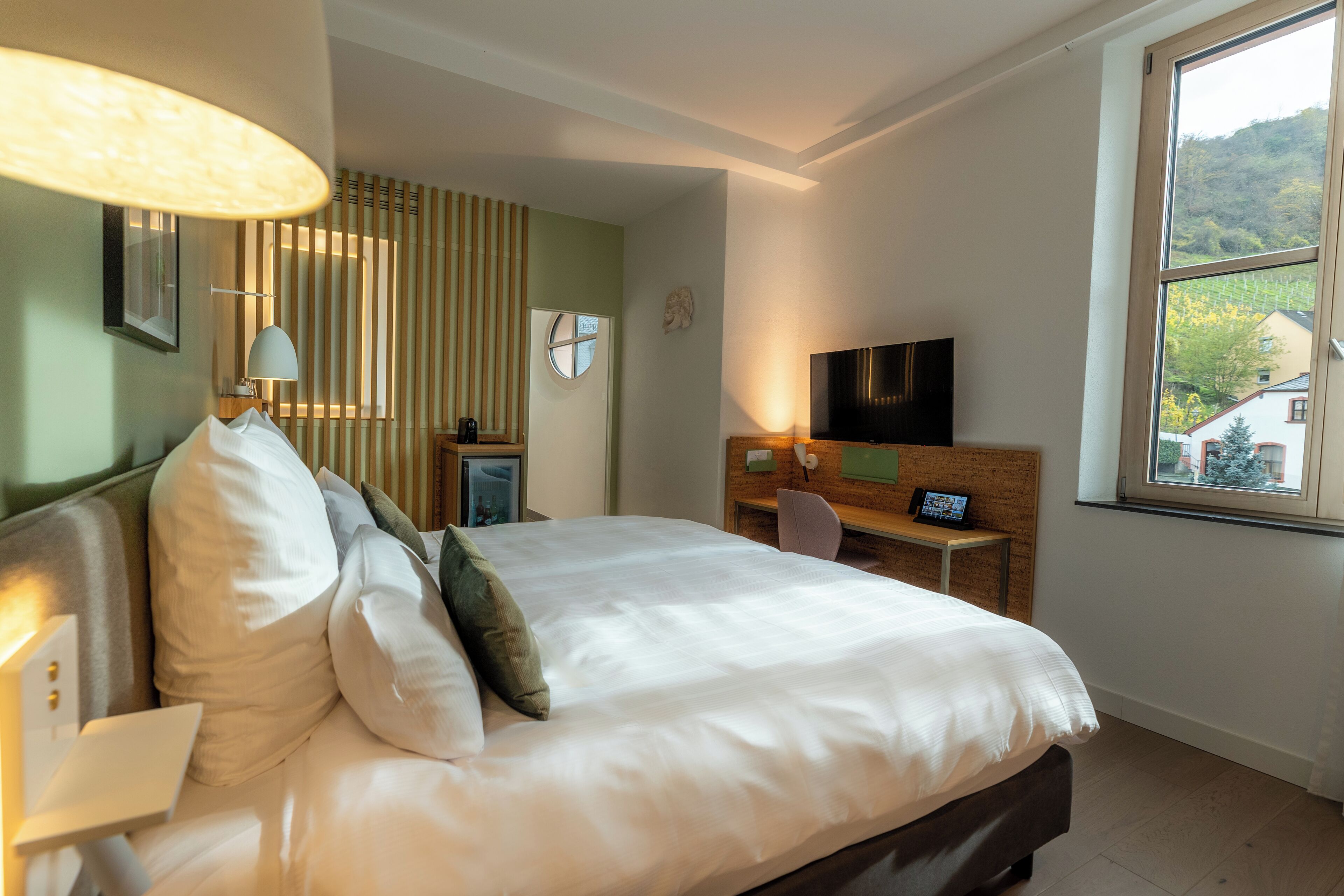 A hotel room with a bed, a TV, a desk, and a window with a view of greenery.