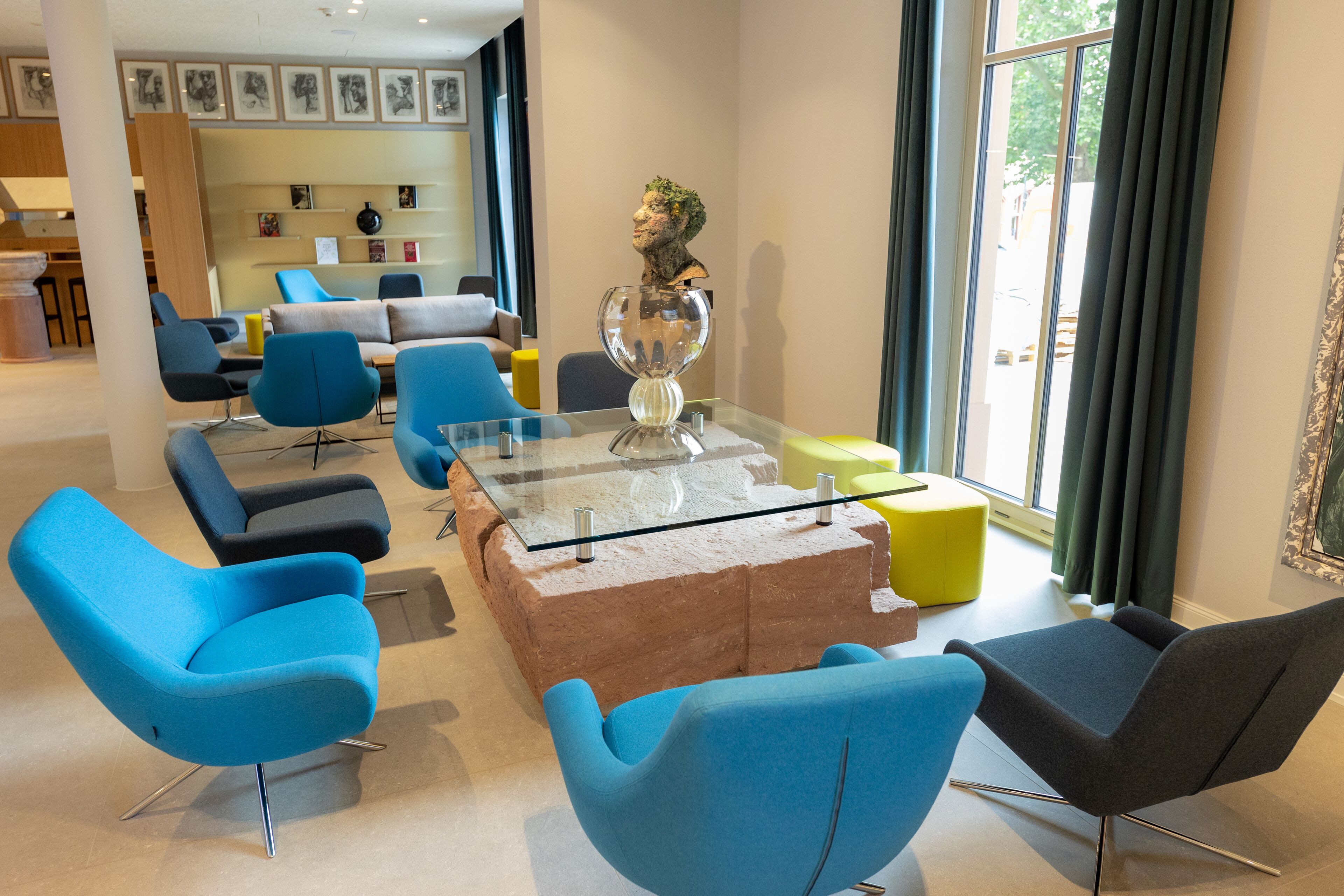 A modern lounge area with blue and black chairs, a glass-top coffee table, and a large window with curtains.