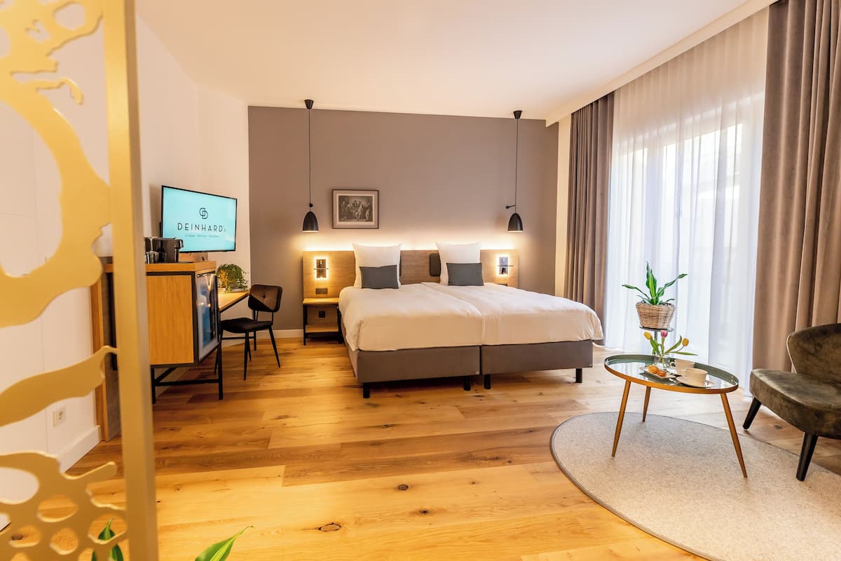 Ein modernes Hotelzimmer mit einem großen Bett, Nachttischlampen, einem Schreibtisch mit Computer, einem Sessel und einem kleinen Tisch mit Pflanze.
