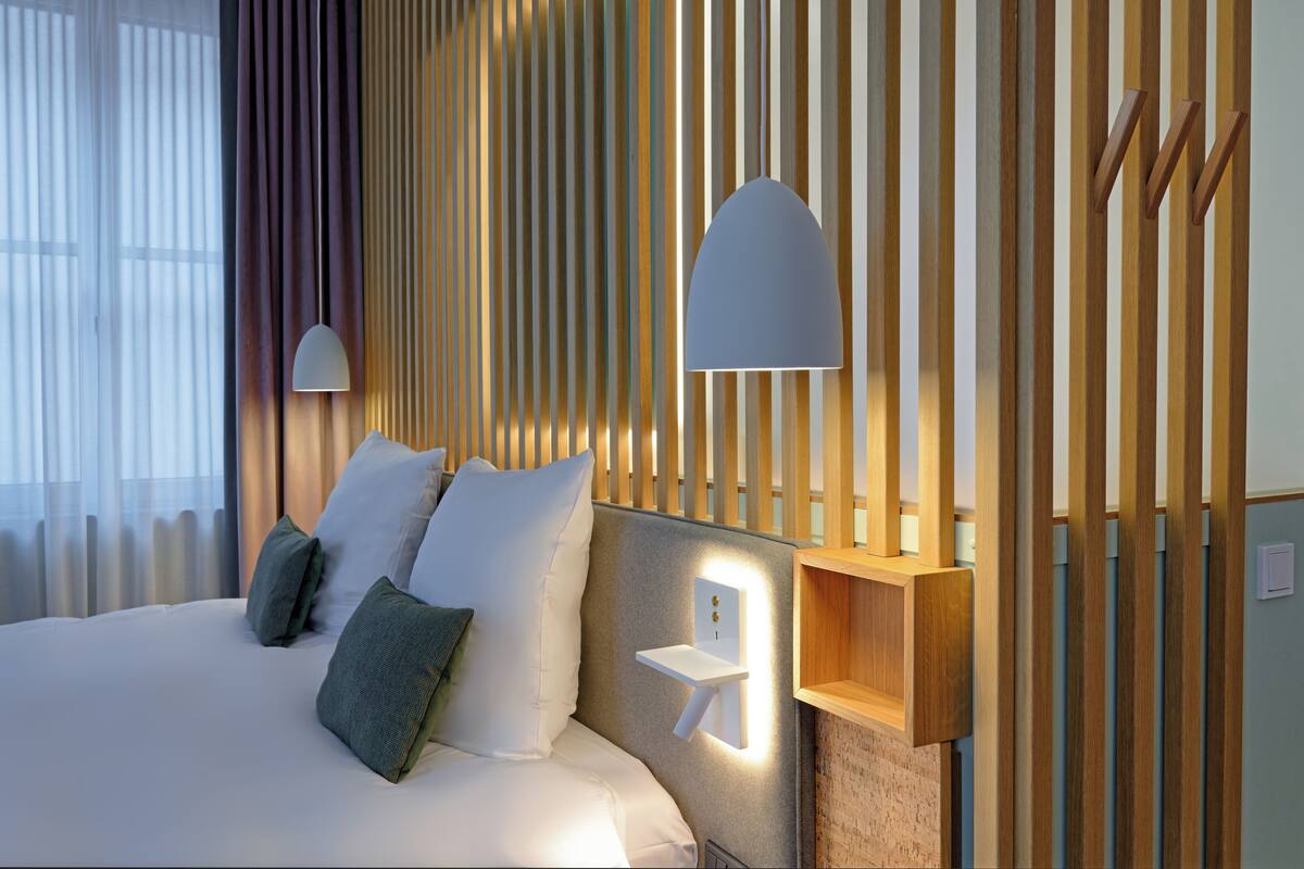 Ein modernes Hotelzimmer mit einem ordentlich bezogenen Bett, einem Nachttisch mit Lampe und einem an der Wand befestigten Regal.