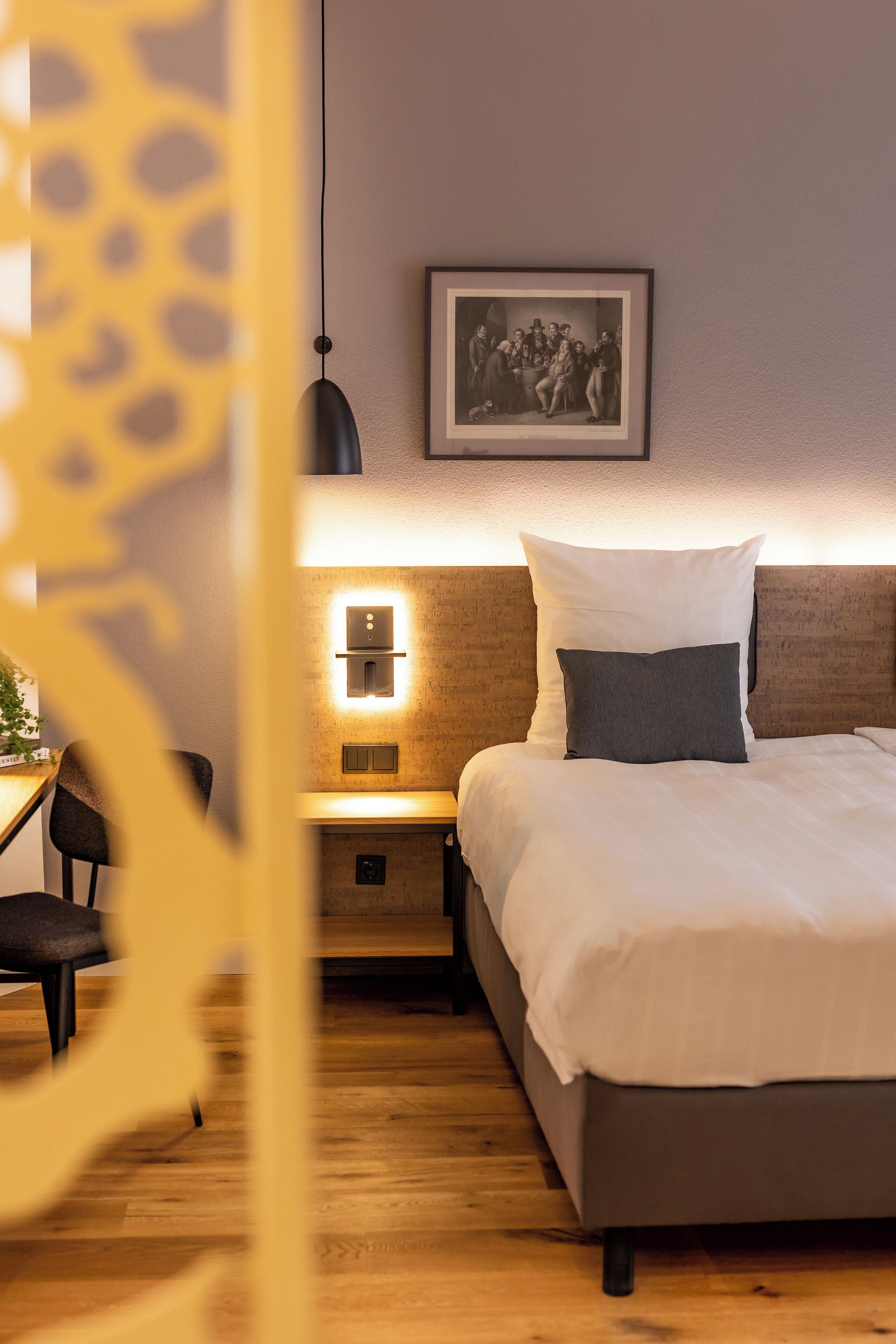 A hotel room with a bed, bedside table, lamp, and a framed picture on the wall.