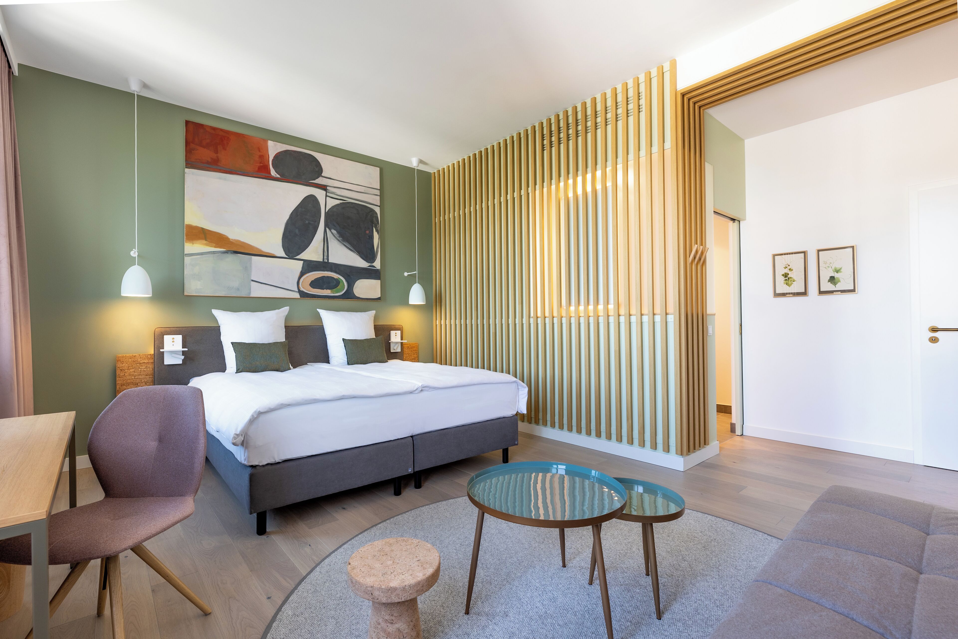 A modern hotel room with a bed, a desk, a chair, two small tables, and a decorative wall piece.