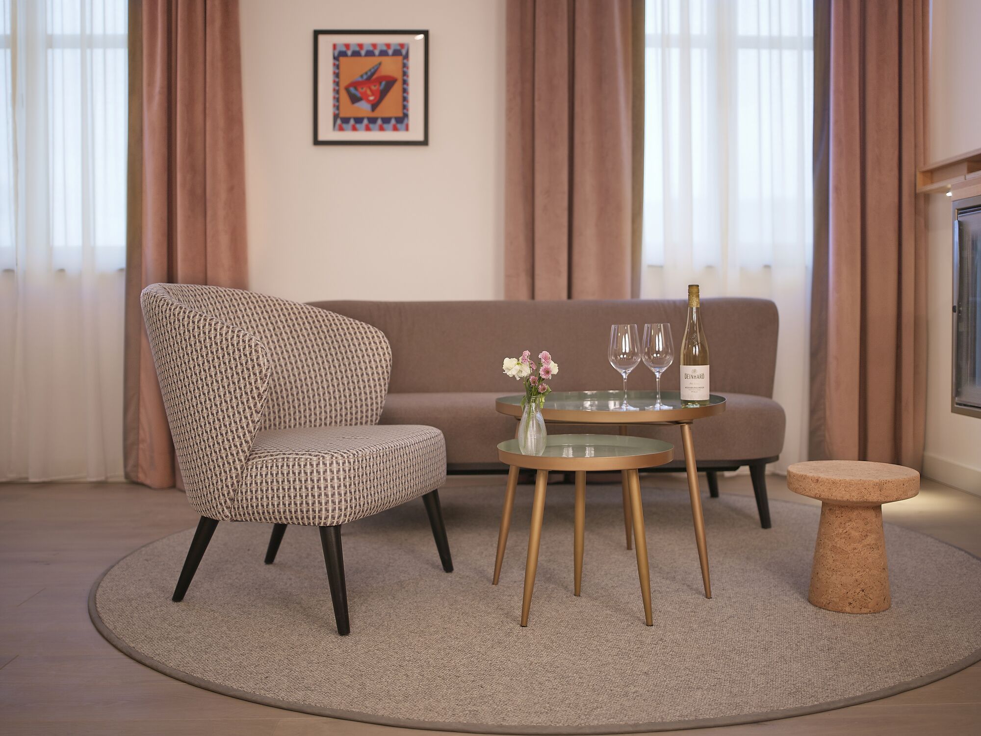 A modern living room with a patterned armchair, a glass coffee table with a bottle of wine and glasses, a small side table, and a framed artwork on the wall.