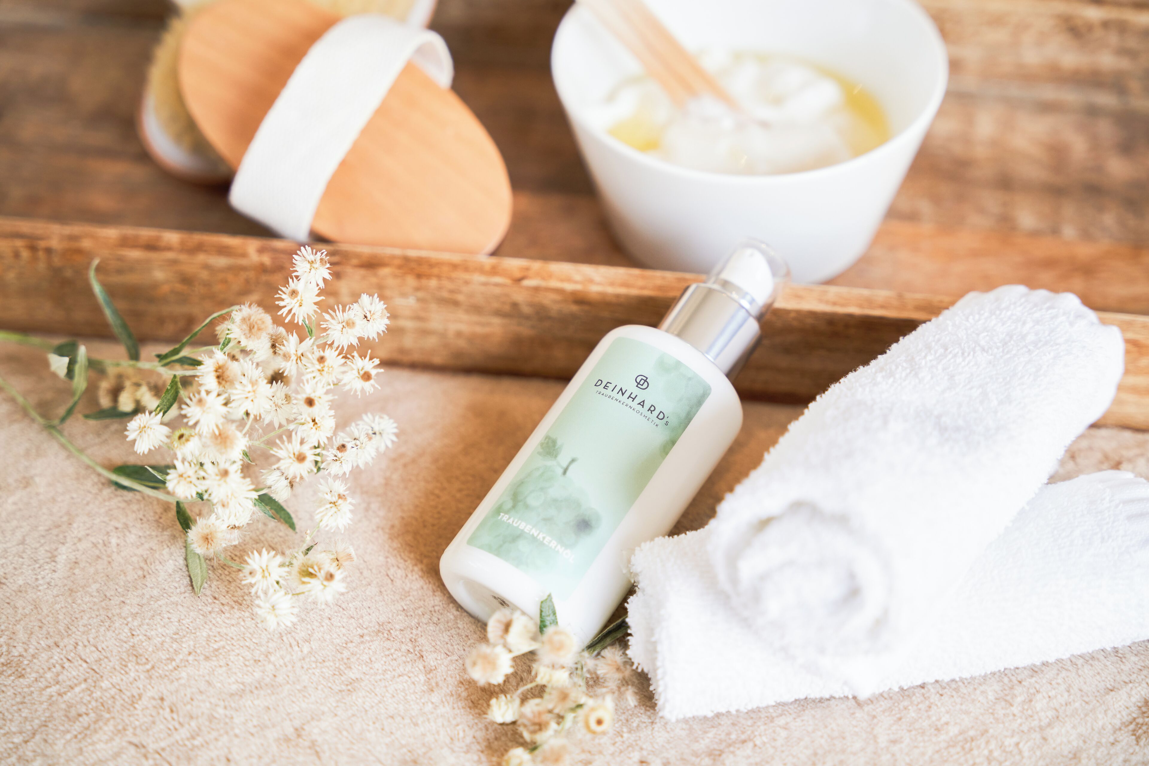 Een fles Deinhard body lotion, een witte handdoek en een kom room op een houten ondergrond.