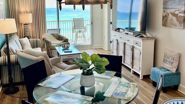 A living room with a glass dining table, a TV, and a view of the ocean. - Majestic Tower 2-502 with RESERVED PARKING SPACE (Panama City Beach)