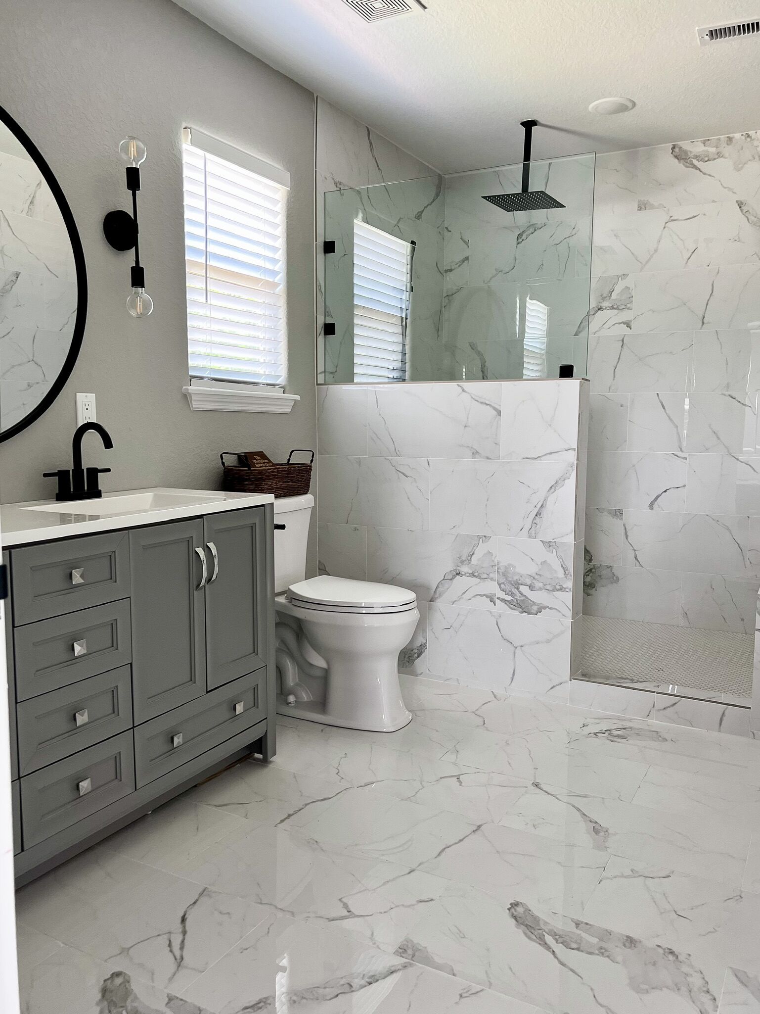 Une salle de bain moderne avec un sol en marbre, des toilettes blanches, un meuble-lavabo gris doté de tiroirs, un miroir rond et une cabine de douche en verre.