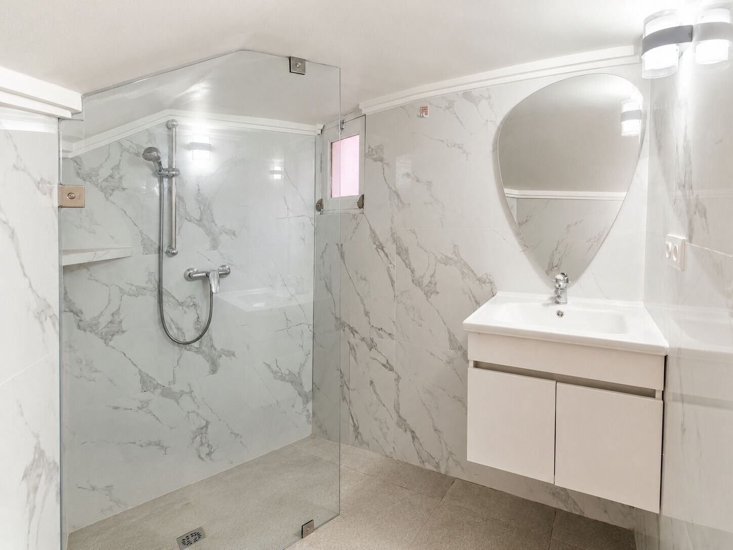 A modern bathroom with a glass-enclosed shower, a marble-tiled wall, a white sink with a single mirror, and a cabinet with drawers.