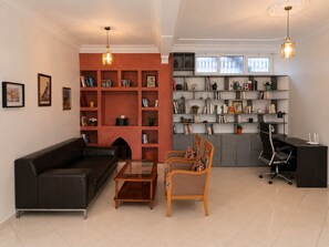 A modern living room with a black sofa, a wooden coffee table, a patterned armchair, and a desk with a black office chair. There are two bookshelves filled with books and decorative items, and a large window with blinds.