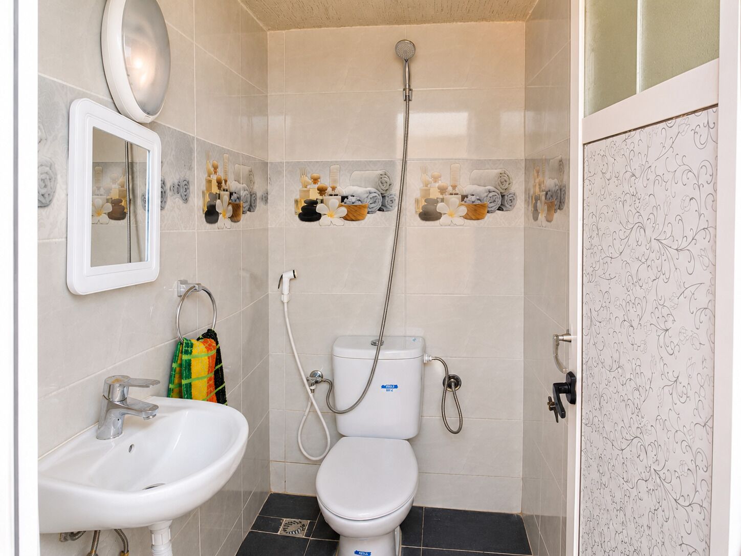 Une salle de bain avec un lavabo, des toilettes et une douche, le tout de couleur blanche. Un miroir est placé au-dessus du lavabo et les murs sont décorés de carreaux.