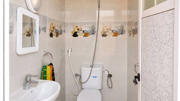 Une salle de bain avec un lavabo, des toilettes et une douche, le tout de couleur blanche. Un miroir est placé au-dessus du lavabo et les murs sont décorés de carreaux.