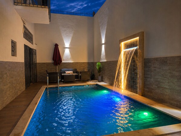 An indoor swimming pool with a waterfall feature, seating area, and a view of the sky.