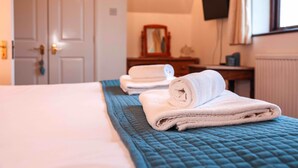 Een net opgemaakt bed met een blauwe deken, een stapel opgevouwen handdoeken en een houten nachtkastje met een spiegel en een klein tafeltje.