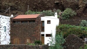 A building with a stone facade and a red-tiled roof, surrounded by greenery and a rocky hillside. - Renovated stone house 6 minutes from Valverde (Mocanal)