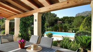 En udendørs patio med udsigt til pool, et trætag, siddepladser og frodig vegetation.