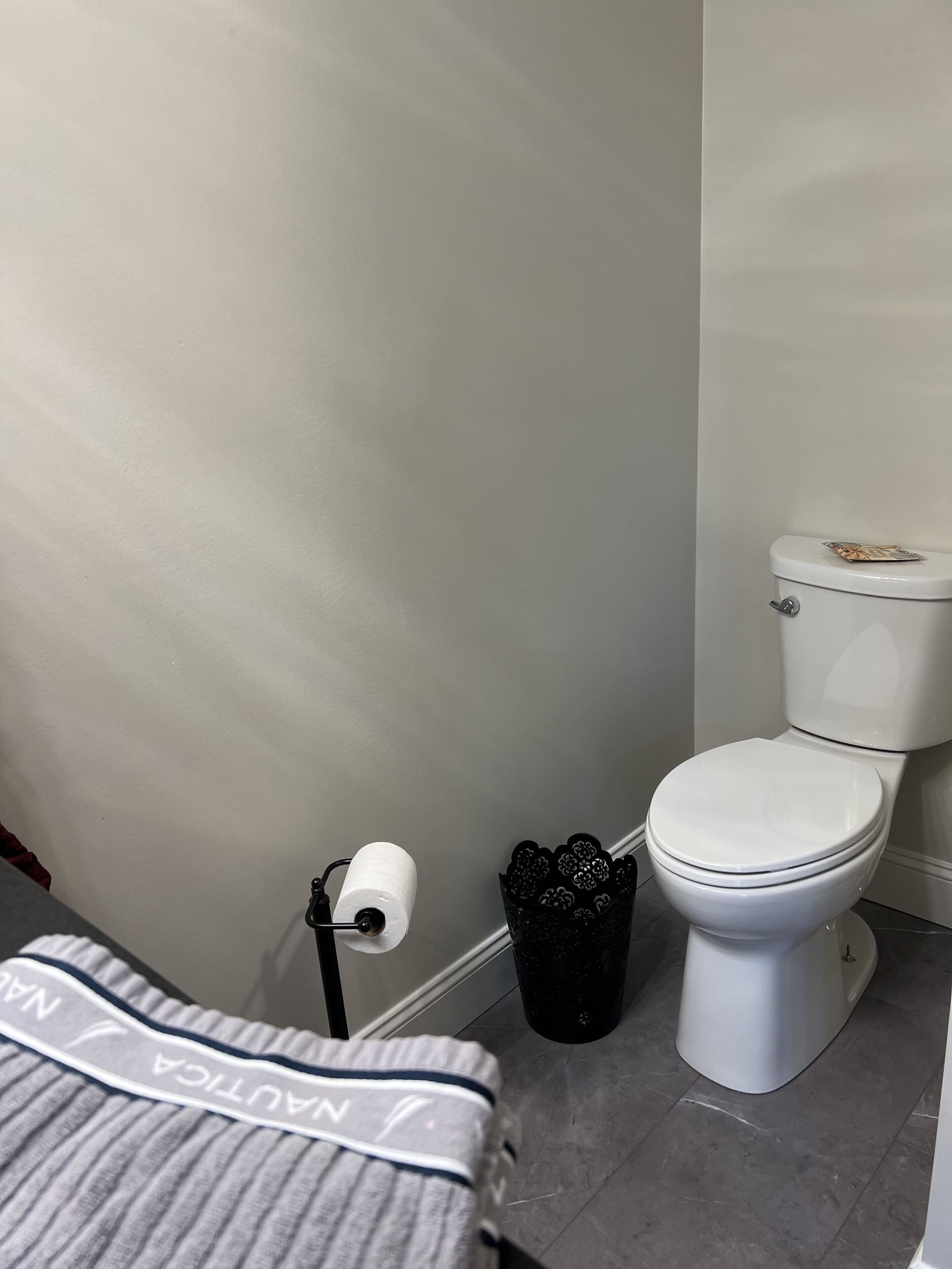 Une salle de bain avec des toilettes, une poubelle noire et un rouleau de papier toilette par terre.
