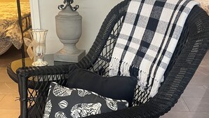 A black wicker chair with a checkered throw blanket and patterned cushions, placed in a room with a tiled floor and a potted plant in the background.