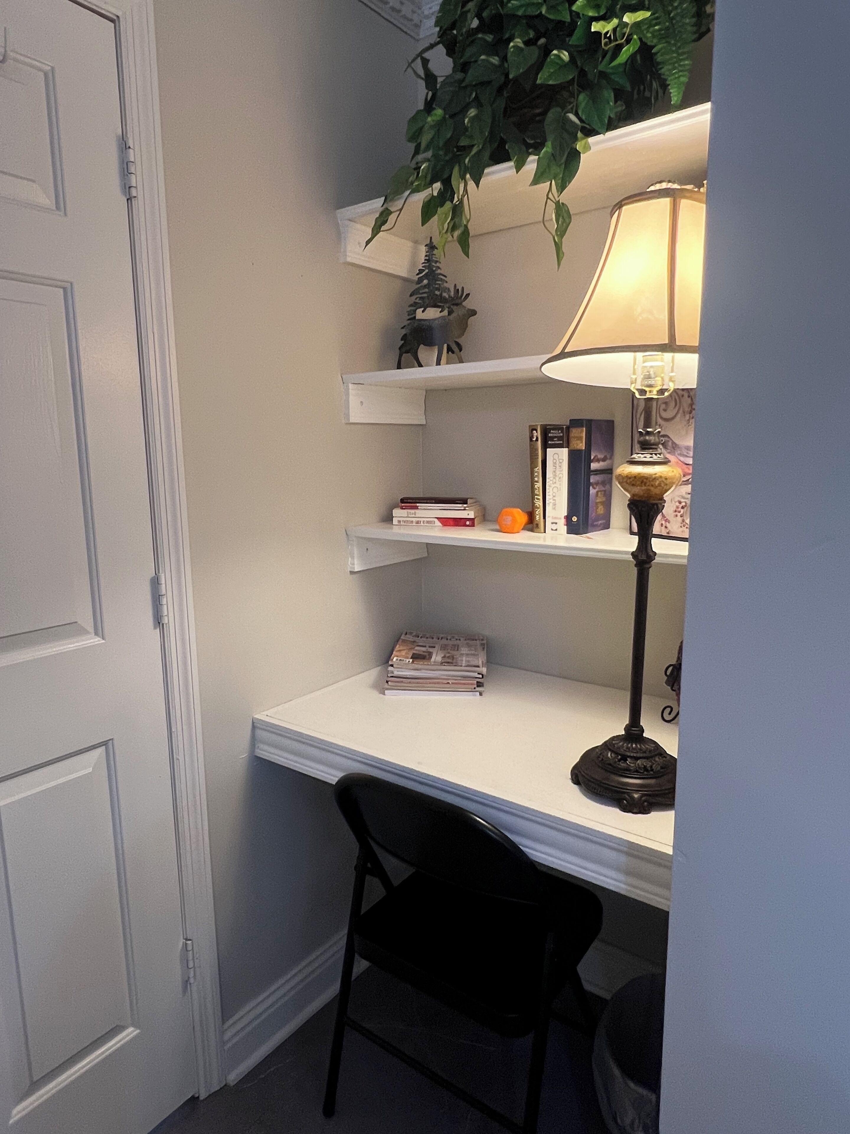 Un petit bureau blanc, avec une chaise noire, une lampe et une plante posée sur une étagère.