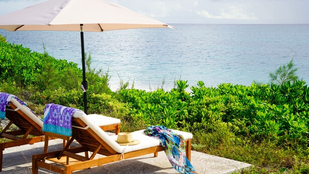 A seaside lounge with two wooden lounge chairs, a large umbrella, and a view of the ocean. - SPECTACULAR OCEAN VIEWS 🌊Kayak, Starlink, Generator, Beach, Pickleball (Crossing Rocks)