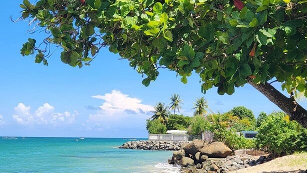 A beach with clear turquoise water, a sandy shore, and a rocky outcrop with a house in the background.