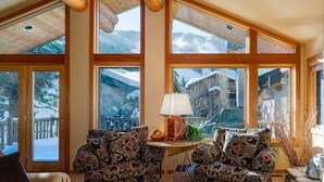 Ein gemütliches Zimmer mit großen Fenstern, zwei gemusterten Sesseln, einem kleinen Tisch mit Lampe und Blick auf verschneite Berge.