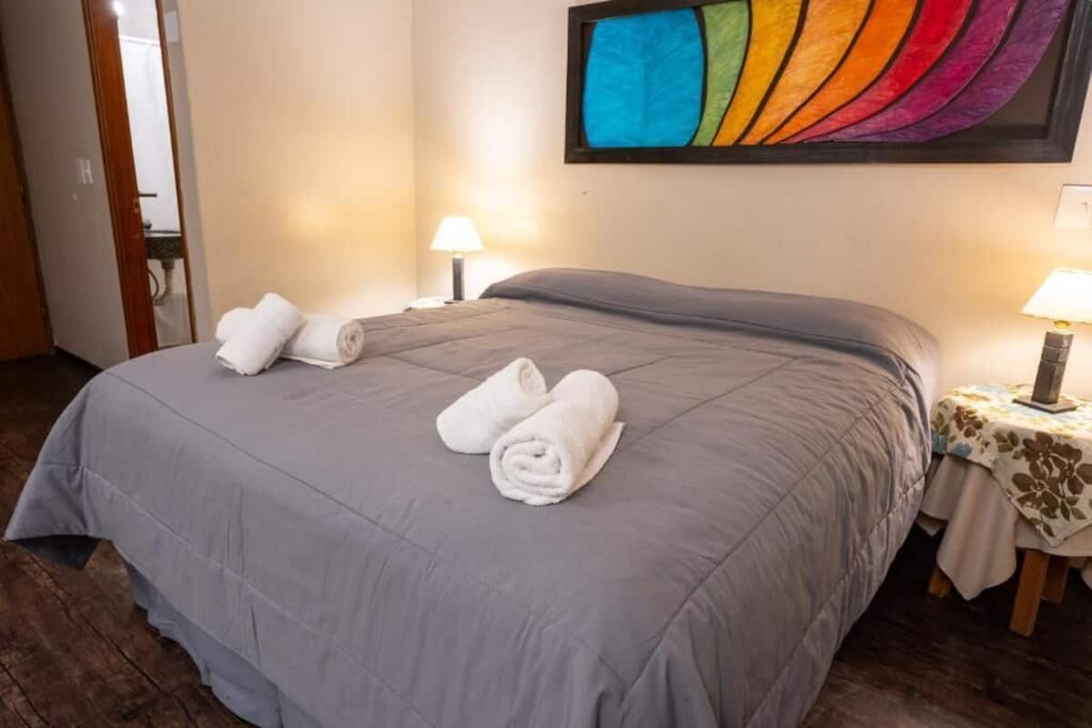 A neatly made bed with a grey bedspread and rolled white towels, a colorful abstract painting on the wall, and a small table with a lamp and a floral tablecloth.