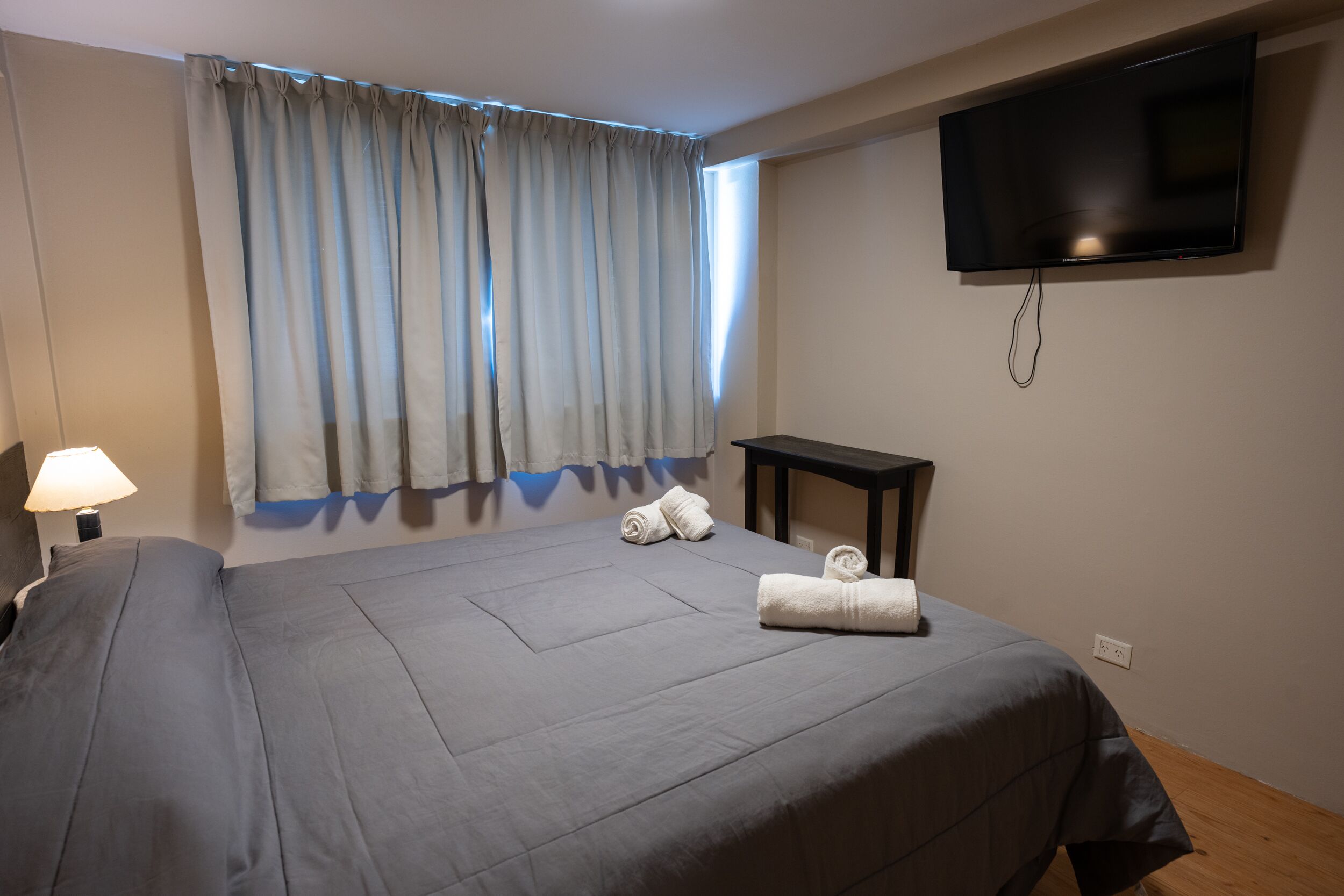 Hotellihuone, jossa on suuri sänky, seinälle asennettu televisio, yöpöytä ja lamppu.