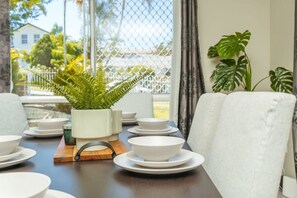 A dining area with a table set for four, a potted fern, and a view through a window with a fence. - Spacious  newly redecorated 5BD Family Home (Eight Mile Plains)