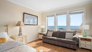 Une chambre à coucher avec un lit, un canapé gris, deux tables de chevet avec des lampes, un tableau encadré au mur et une grande fenêtre donnant sur la plage.