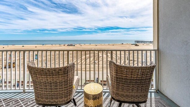 Un balcon agrémenté de fauteuils en osier et d’une table ronde, d’où l’on peut admirer une plage et, au loin, une jetée.