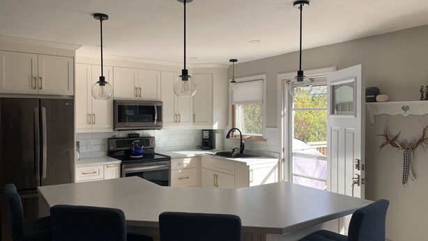 A modern kitchen with white cabinets, a stainless steel refrigerator, a microwave, and a kettle on the stove. There are three black chairs at a white table. - Cozy open concept home, walking distance to Canada Games grounds (Charlottetown)