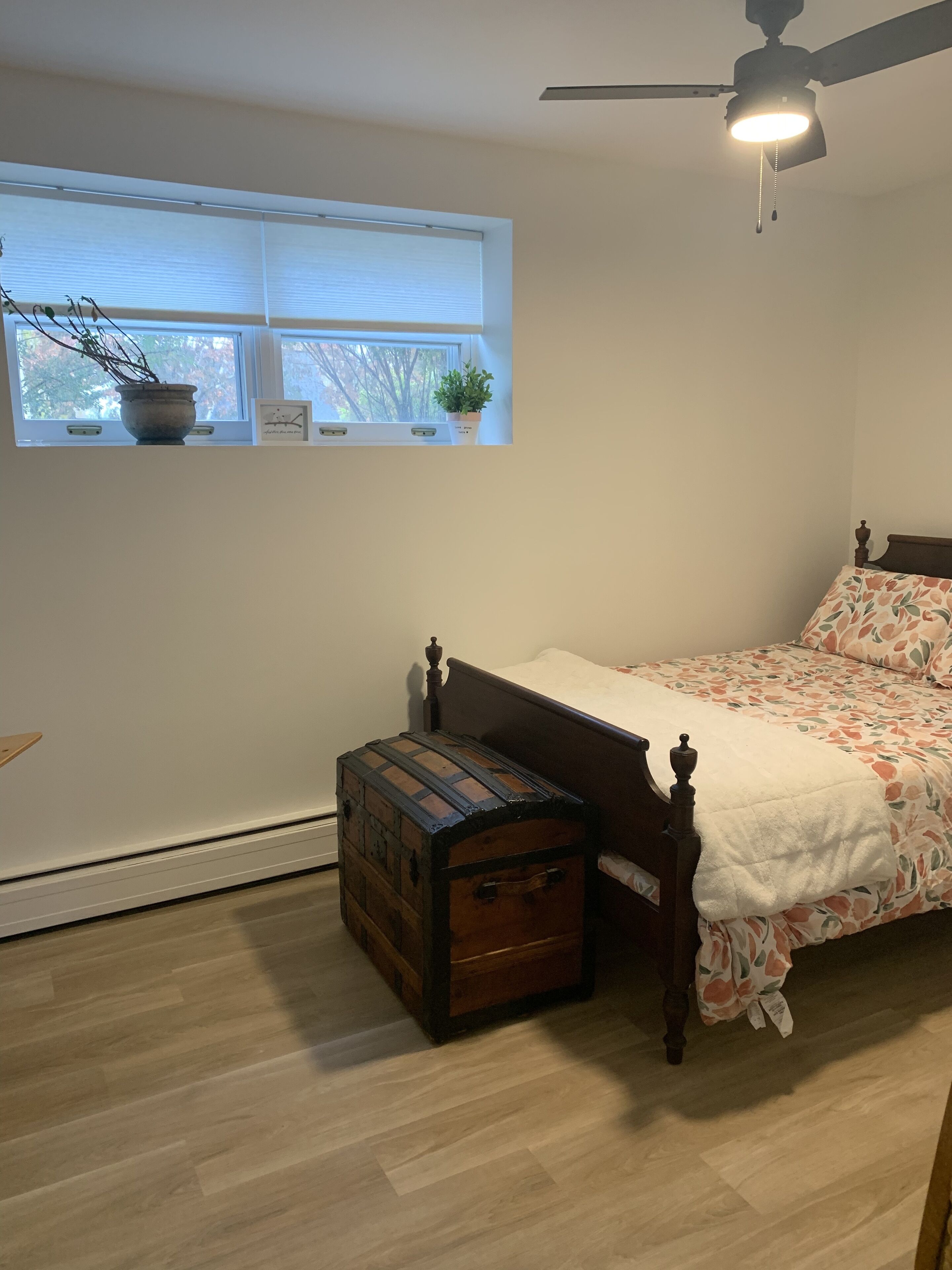Un dormitorio con una cama, un cofre de madera, una ventana con plantas y un ventilador de techo.