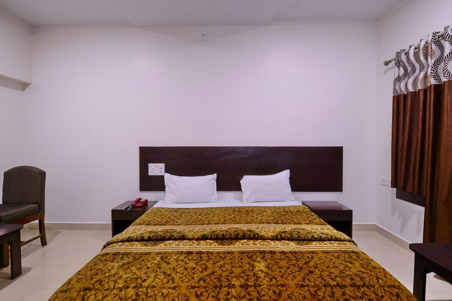 A hotel room with a bed, two white pillows, a brown headboard, a bedside table with a red telephone, a brown chair, and a patterned bedspread.