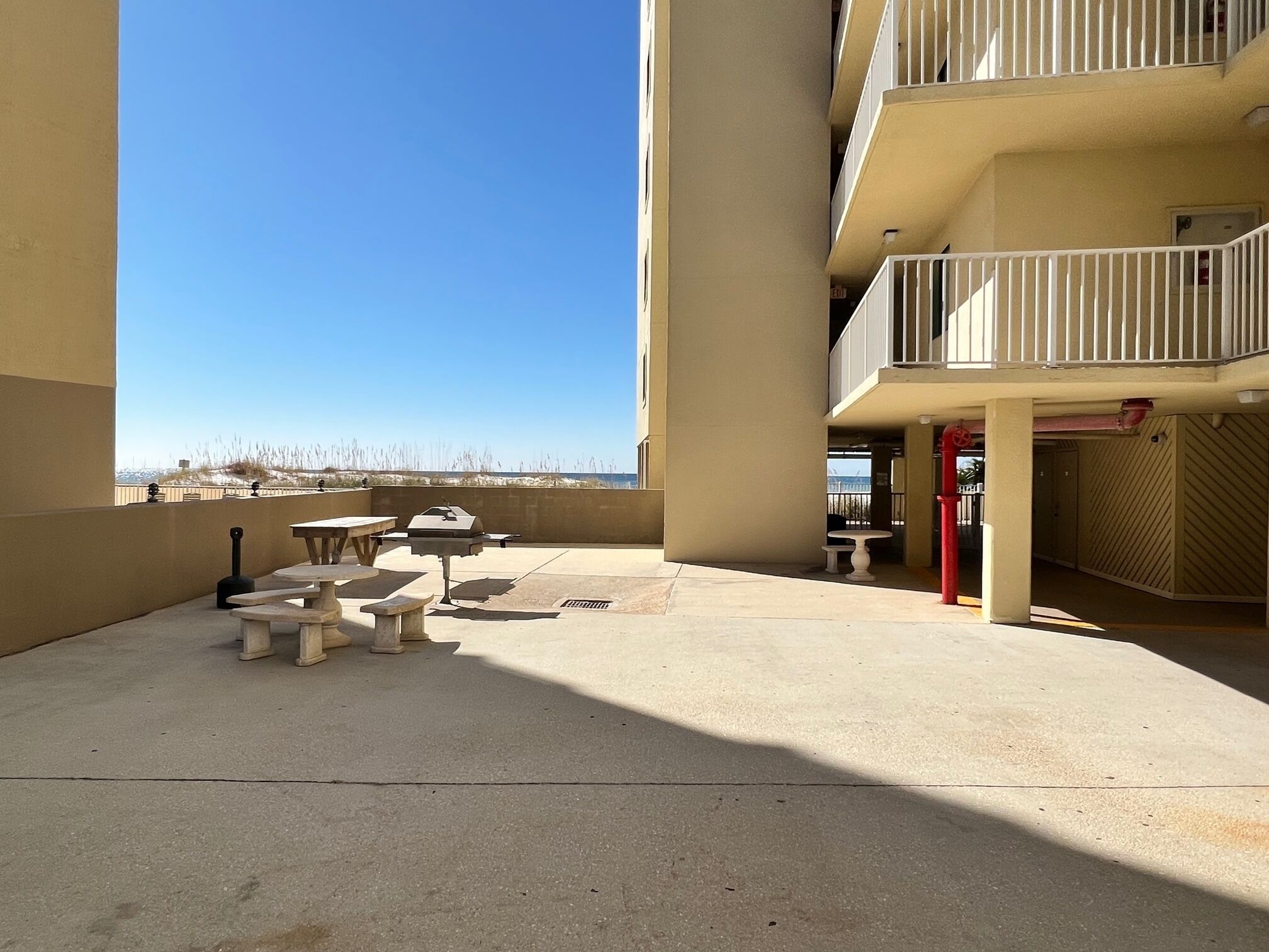 Ein Außenbereich mit einer Terrasse, Beton-Tischen und -Bänken, einem Grill und Blick auf den Strand.